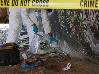 Person in protective gear at a crime scene with 'Do Not Cross' tape and evidence collection.