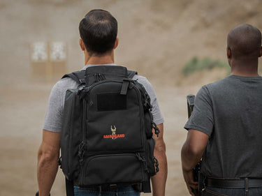 Two men, one with a black backpack featuring a logo, walking on a dirt path.
